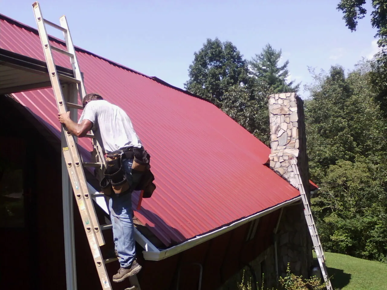 Roofer on a ladder beside a red metal roof and stone chimney — Soffit & Fascia Repair by Roofing Brothers in Galvin